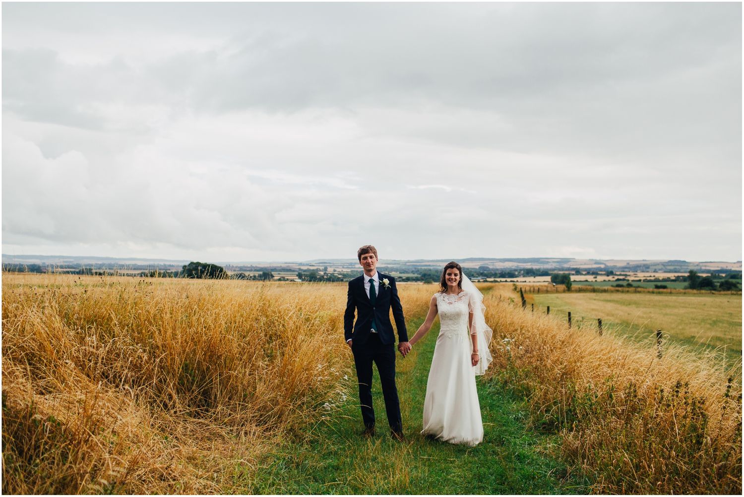 Wedding couple strolling through tall grass with scenic countryside views in background.