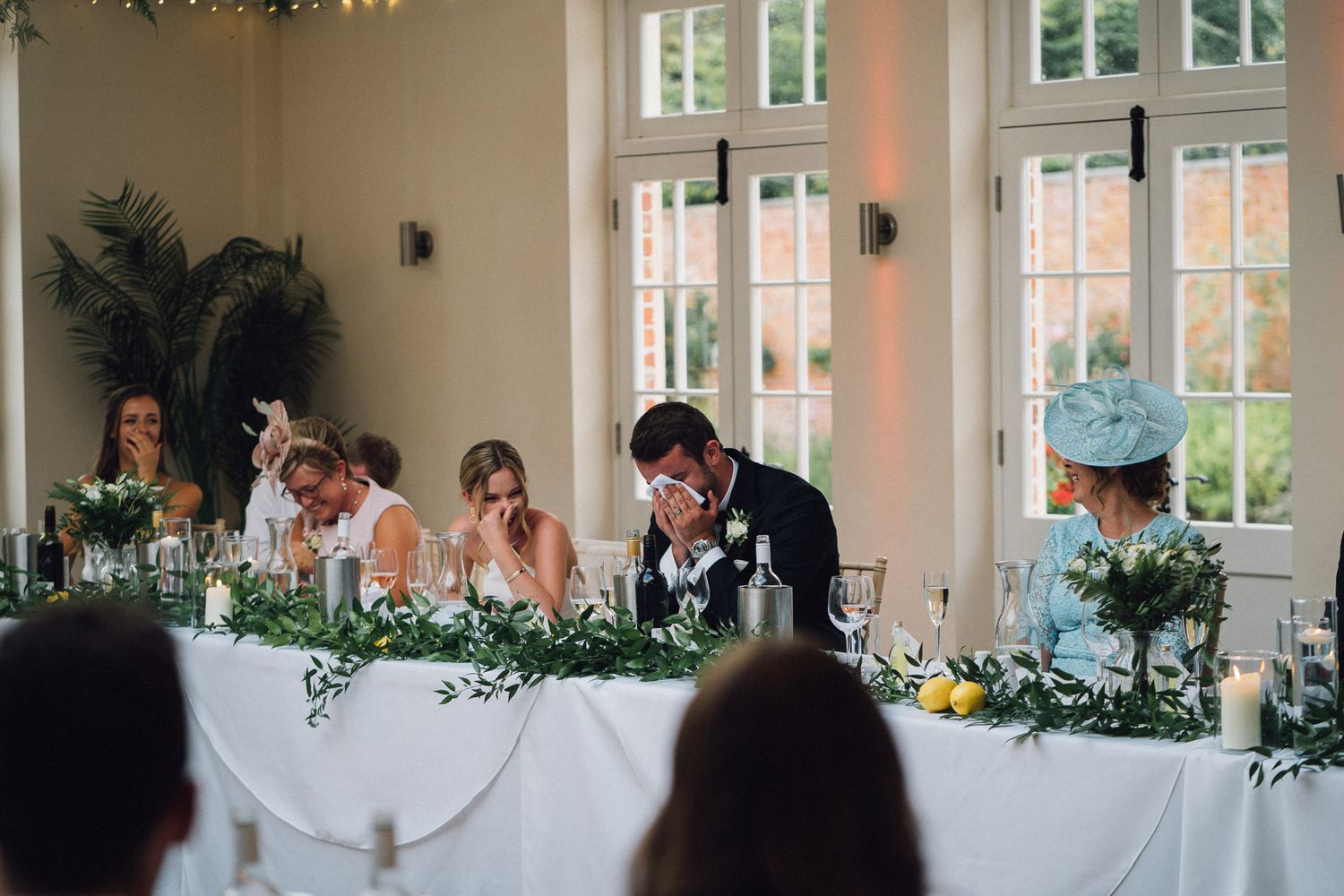 Wedding reception venue decorated with greenery and white linens in bright indoor space.