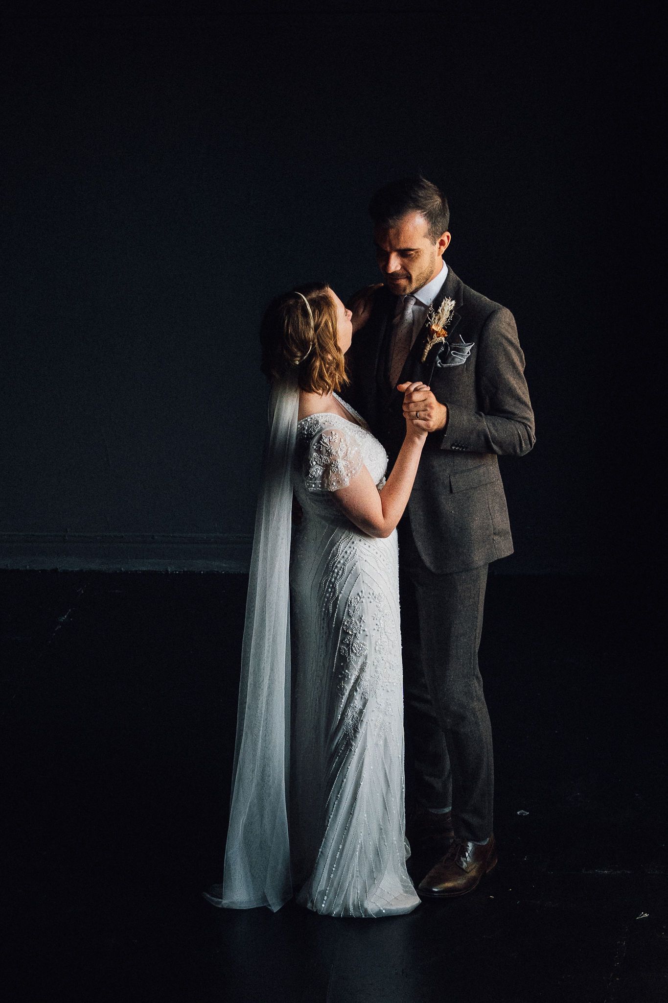 A couple shares an intimate dance moment in a dark room with dramatic lighting and elegant wedding attire.