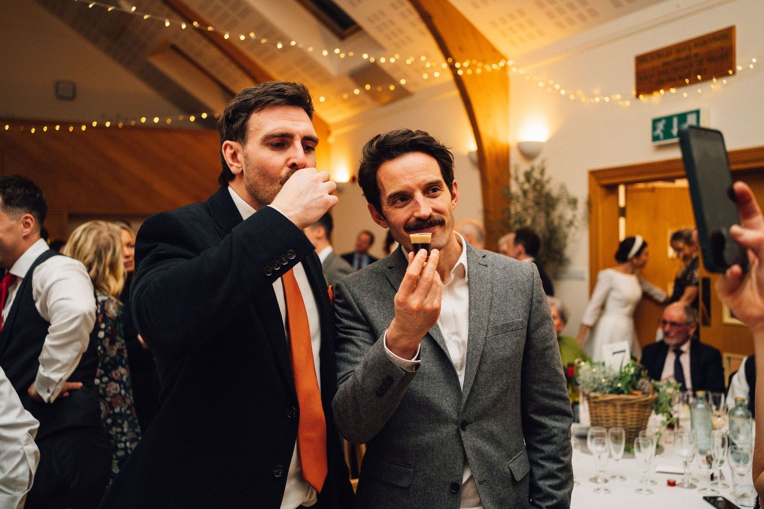 Wedding reception guests mingle and socialize at elegant venue with string lights and vaulted wooden ceiling.