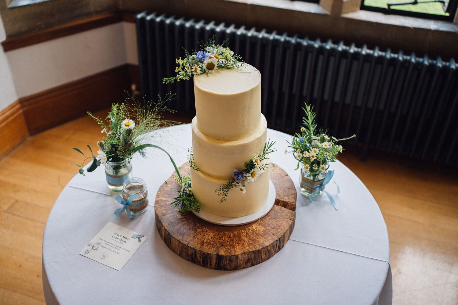 A rustic wedding cake with white frosting sits on a wooden slice platter decorated with fresh greenery and flowers.