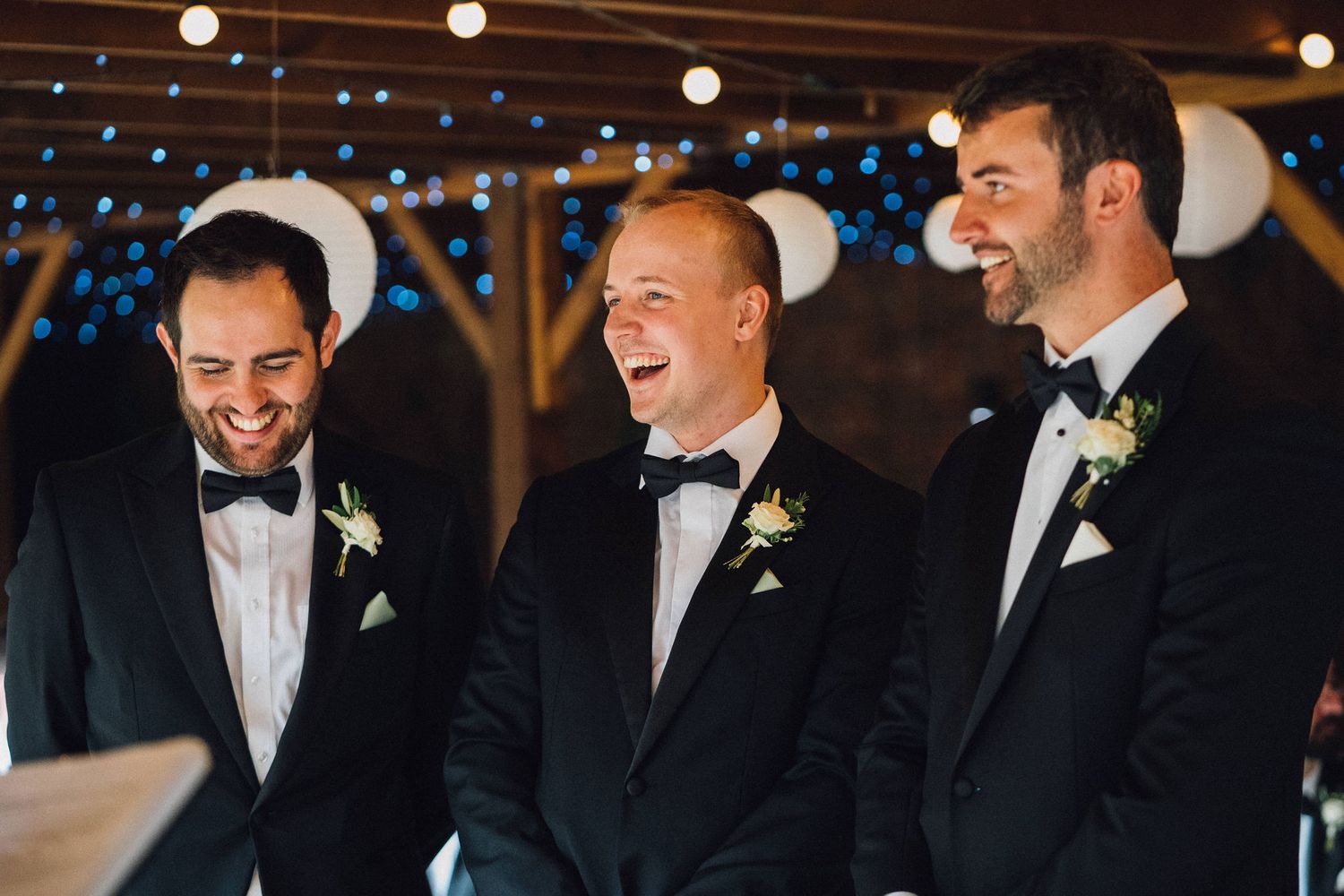Groomsmen in black tuxedos and bowties share candid laughs during a wedding celebration with twinkling lights overhead.