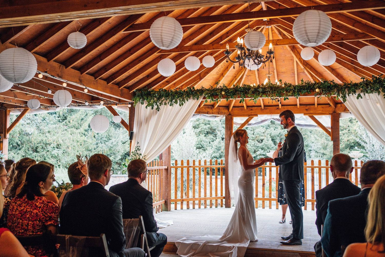 Wedding ceremony taking place under a rustic wooden pavilion decorated with white paper lanterns and flowing fabric.