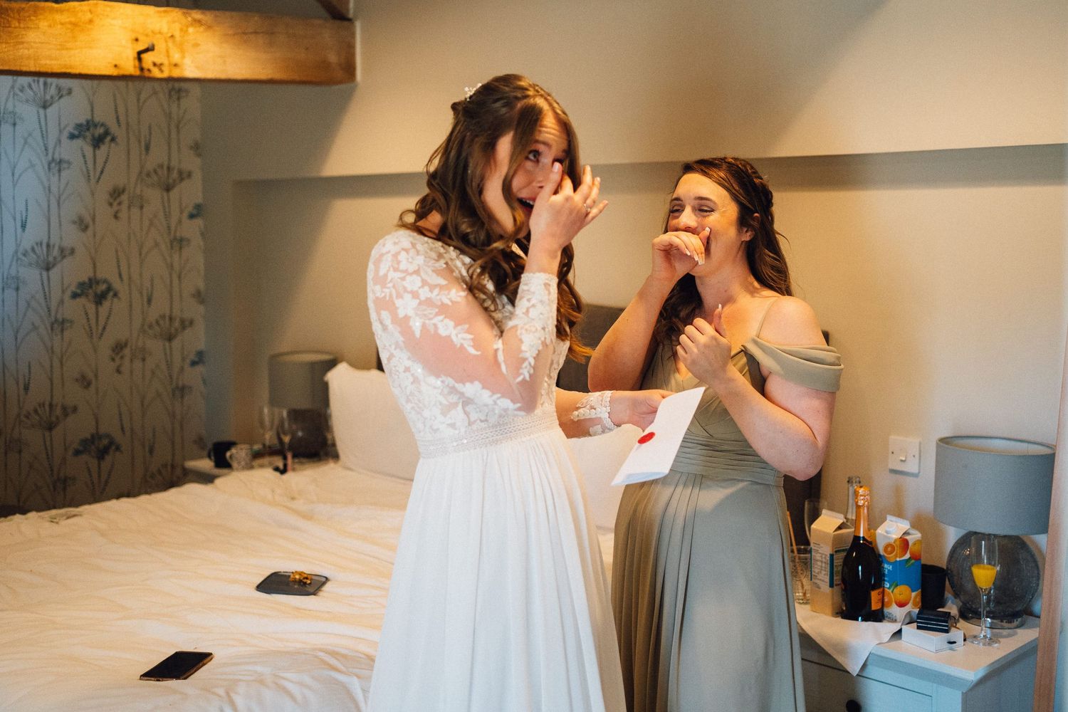 Two women in white dresses share an emotional moment while getting ready in a bright room.