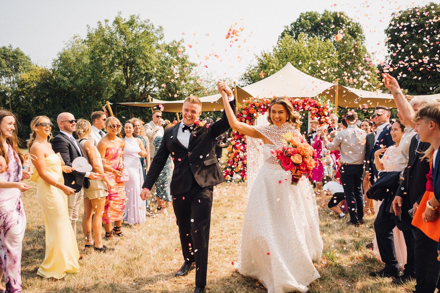 Celebratory scene at an outdoor summer wedding with confetti being tossed in the air under trees.