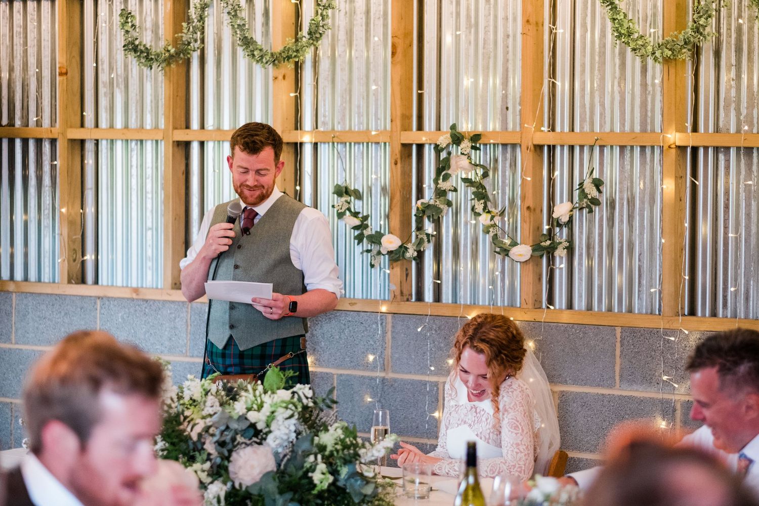 Wedding reception venue decorated with hanging greenery and string lights in a rustic barn setting.