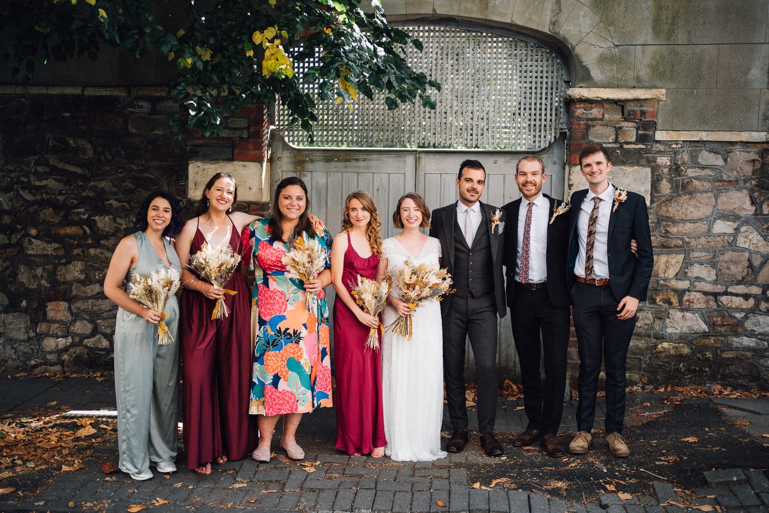 Wedding party in mix of burgundy dresses and dark suits poses together against stone wall backdrop.