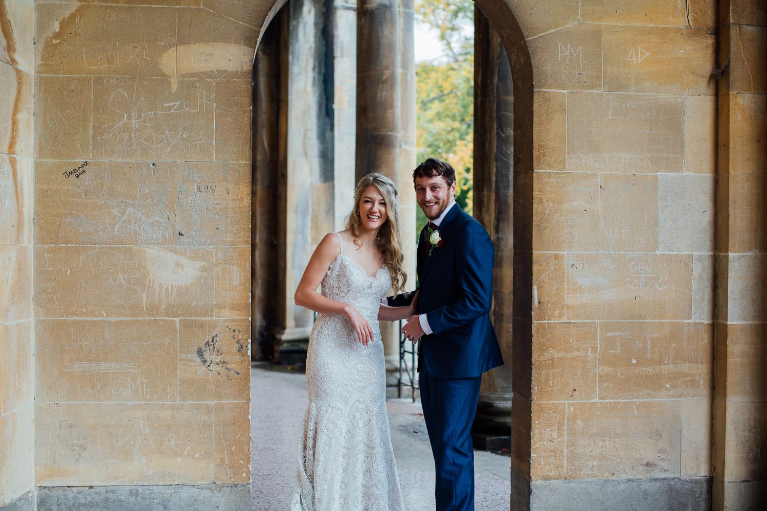Bride in white lace gown and groom in navy suit smiling together under stone archway at elegant wedding venue.