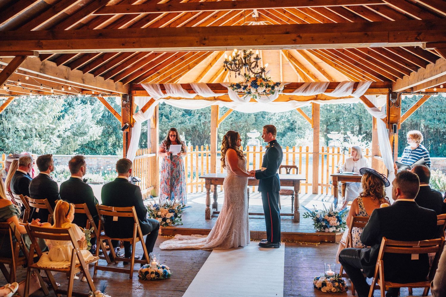 Outdoor wedding ceremony under a wooden gazebo with guests seated, couple exchanging vows before an officiant.