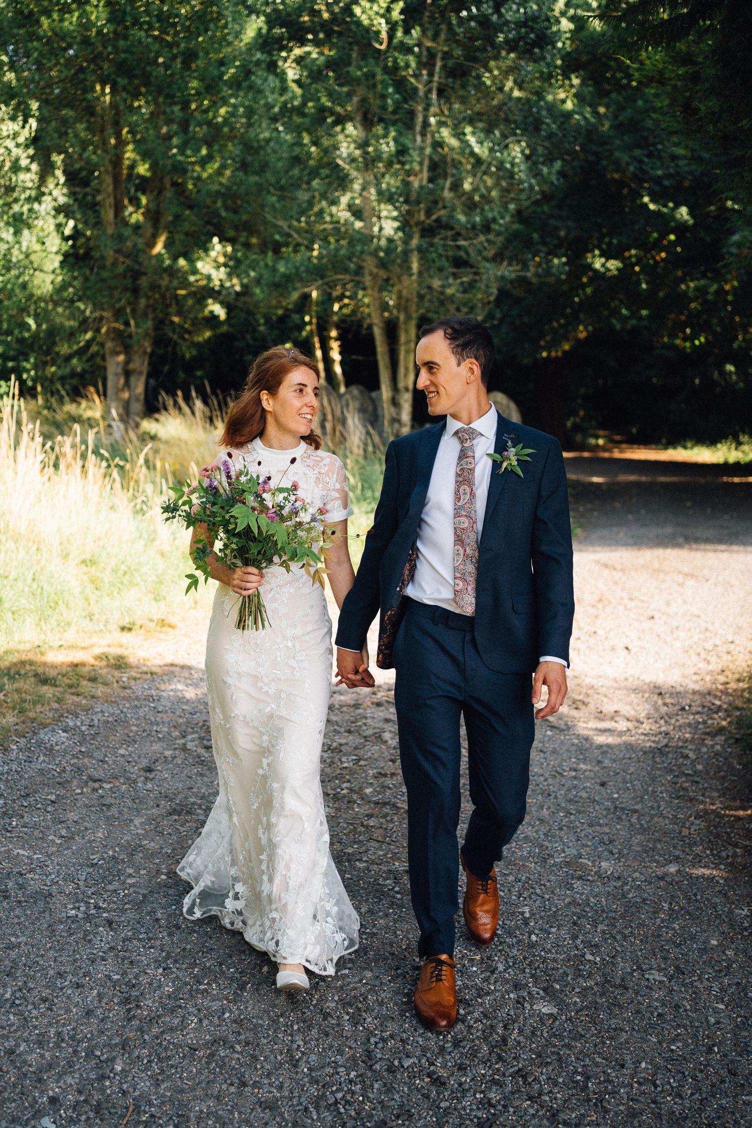 A bride and groom walk hand-in-hand on a gravel path surrounded by lush green trees on their wedding day.