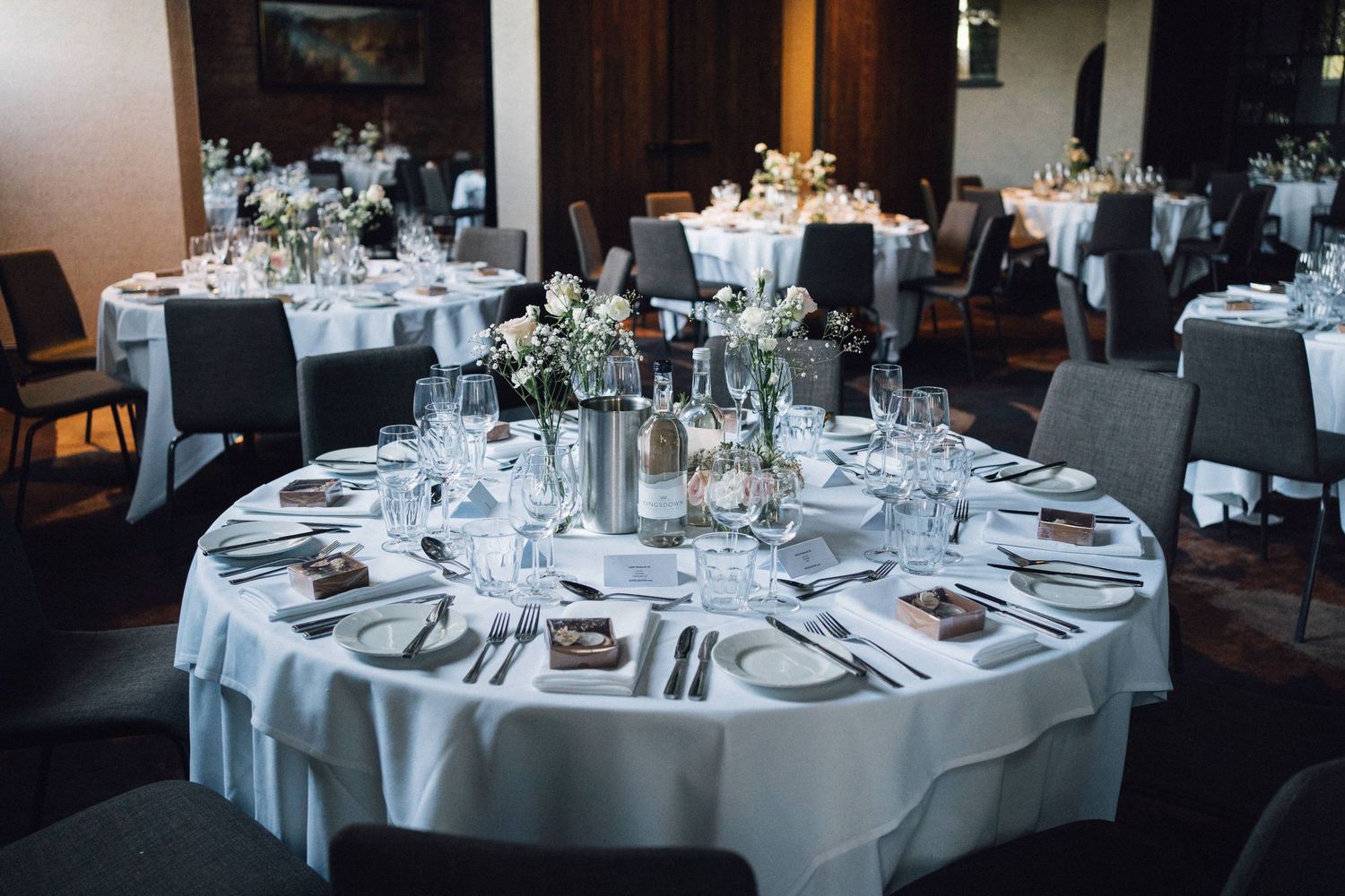 Elegant wedding reception venue with round tables set with white linens, glassware, and floral centerpieces in a dimly lit room.