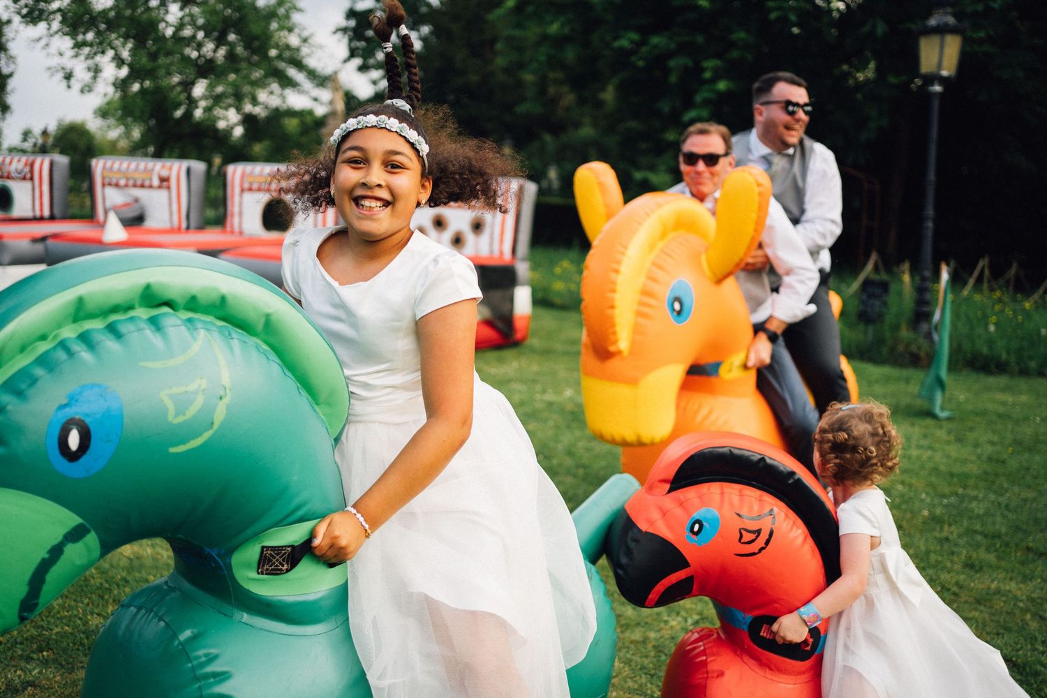 Children in white dresses playing on colorful inflatable animals at outdoor wedding reception on green lawn.
