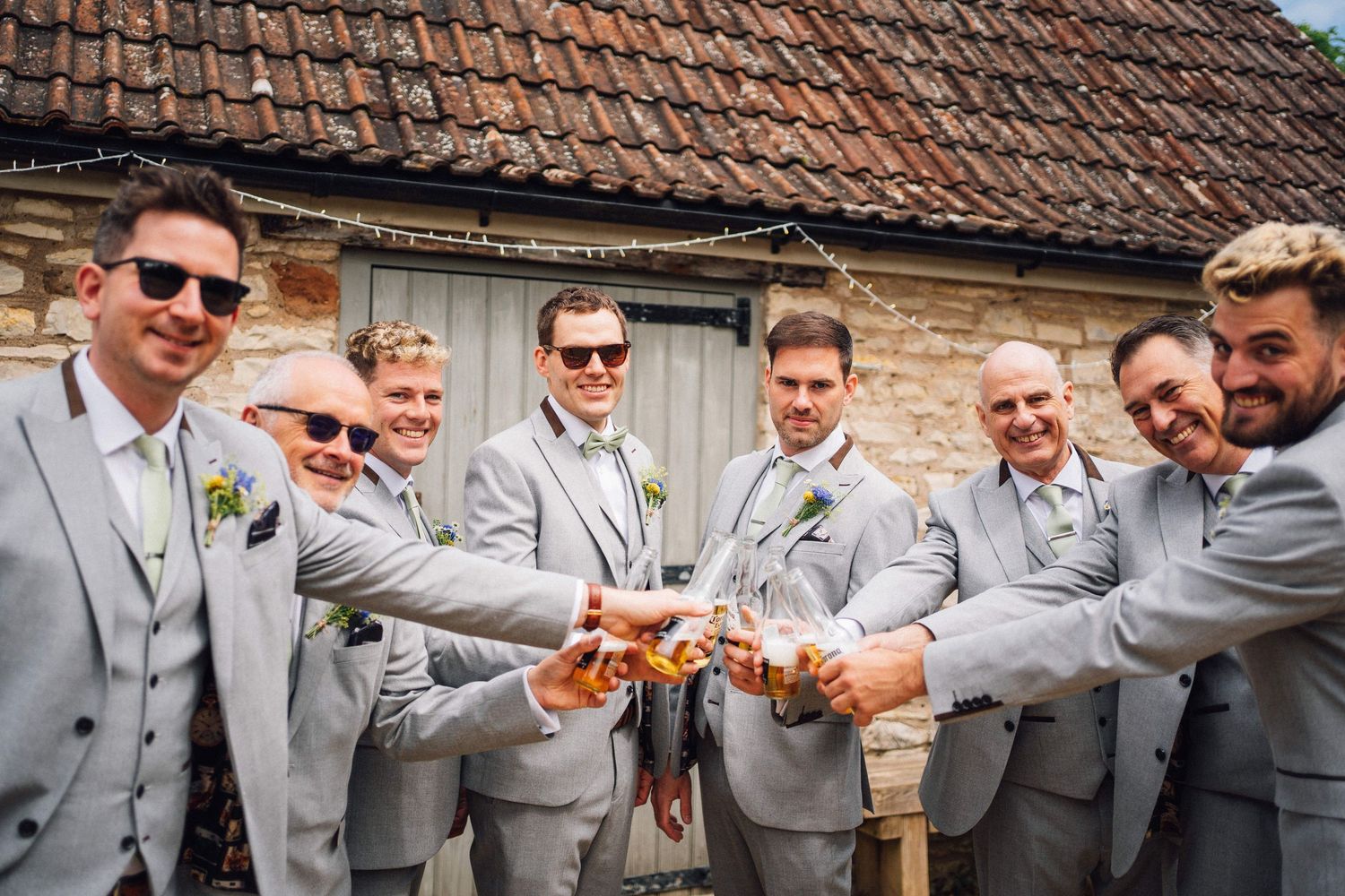 Groomsmen in matching gray suits toasting drinks together outdoors near a rustic stone barn at a wedding celebration.