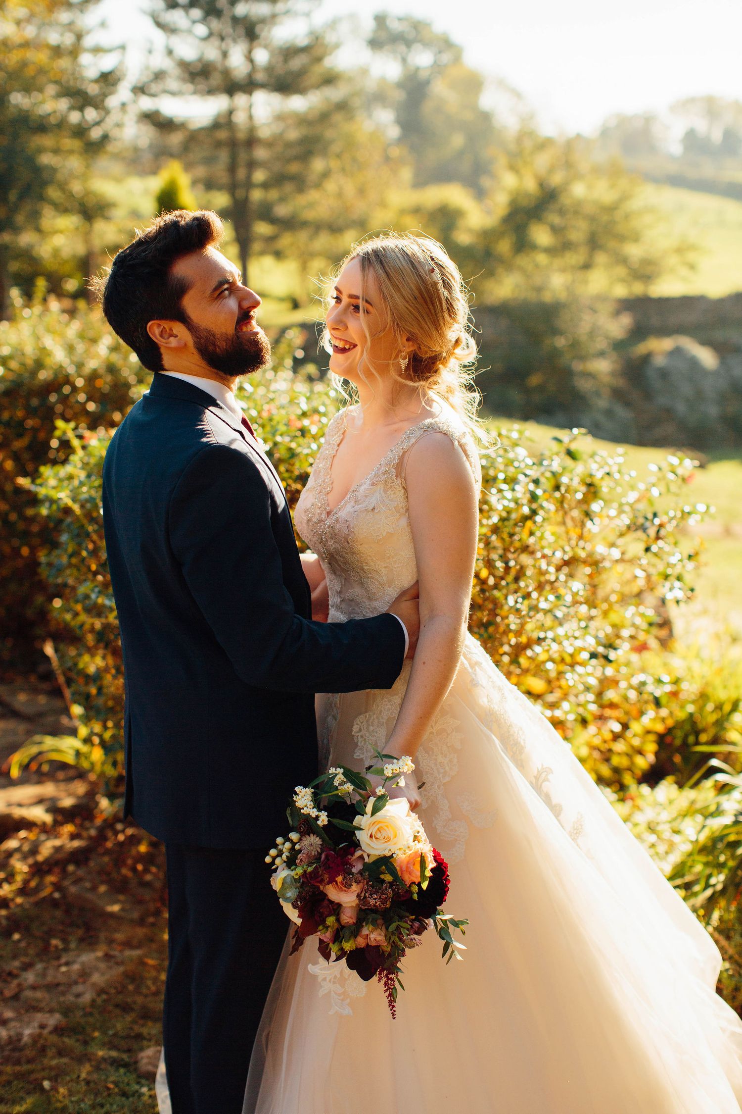 Couple in wedding attire poses in golden sunlight against autumn foliage.