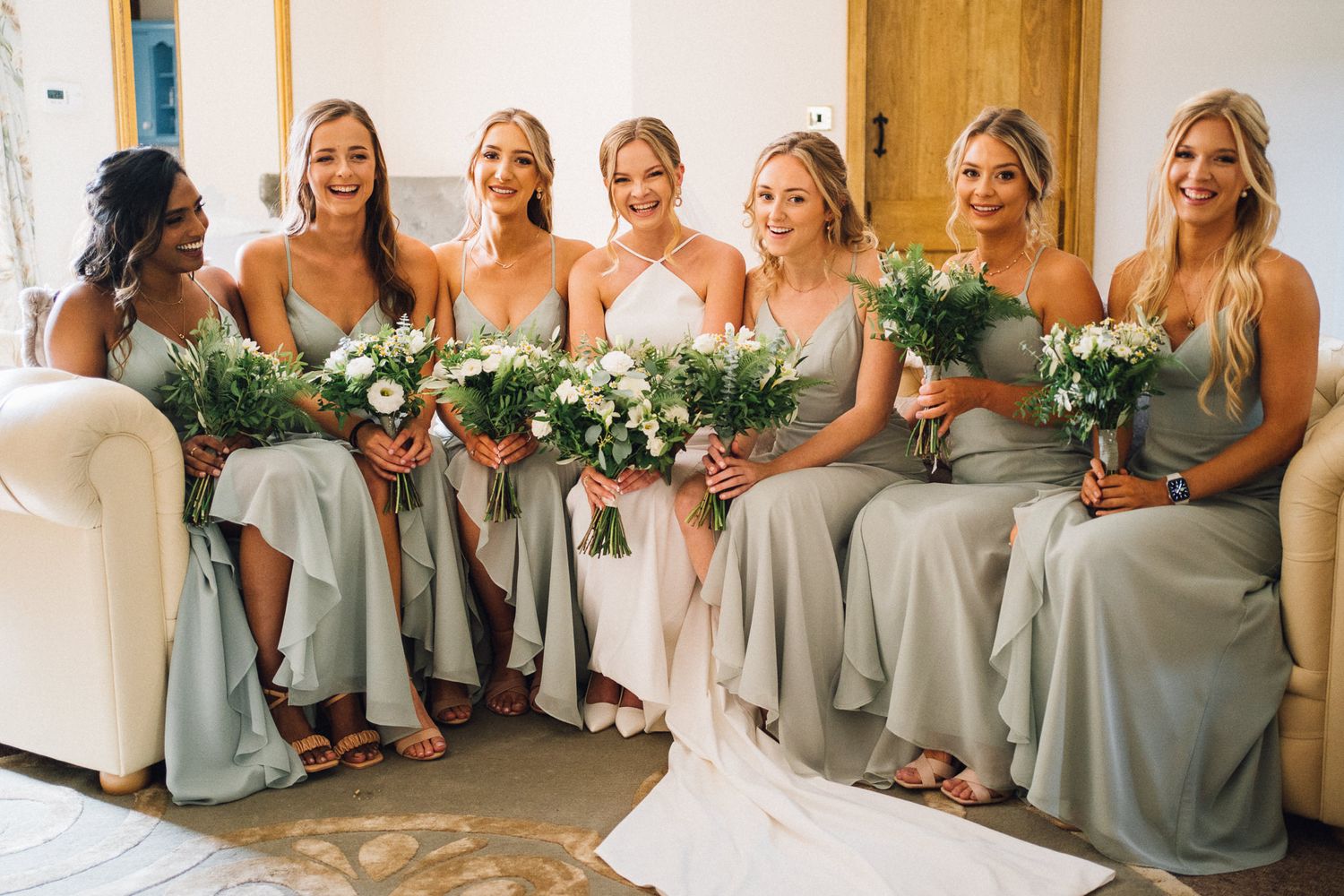 Bridesmaids in sage green dresses pose together holding white floral bouquets at a wedding celebration.