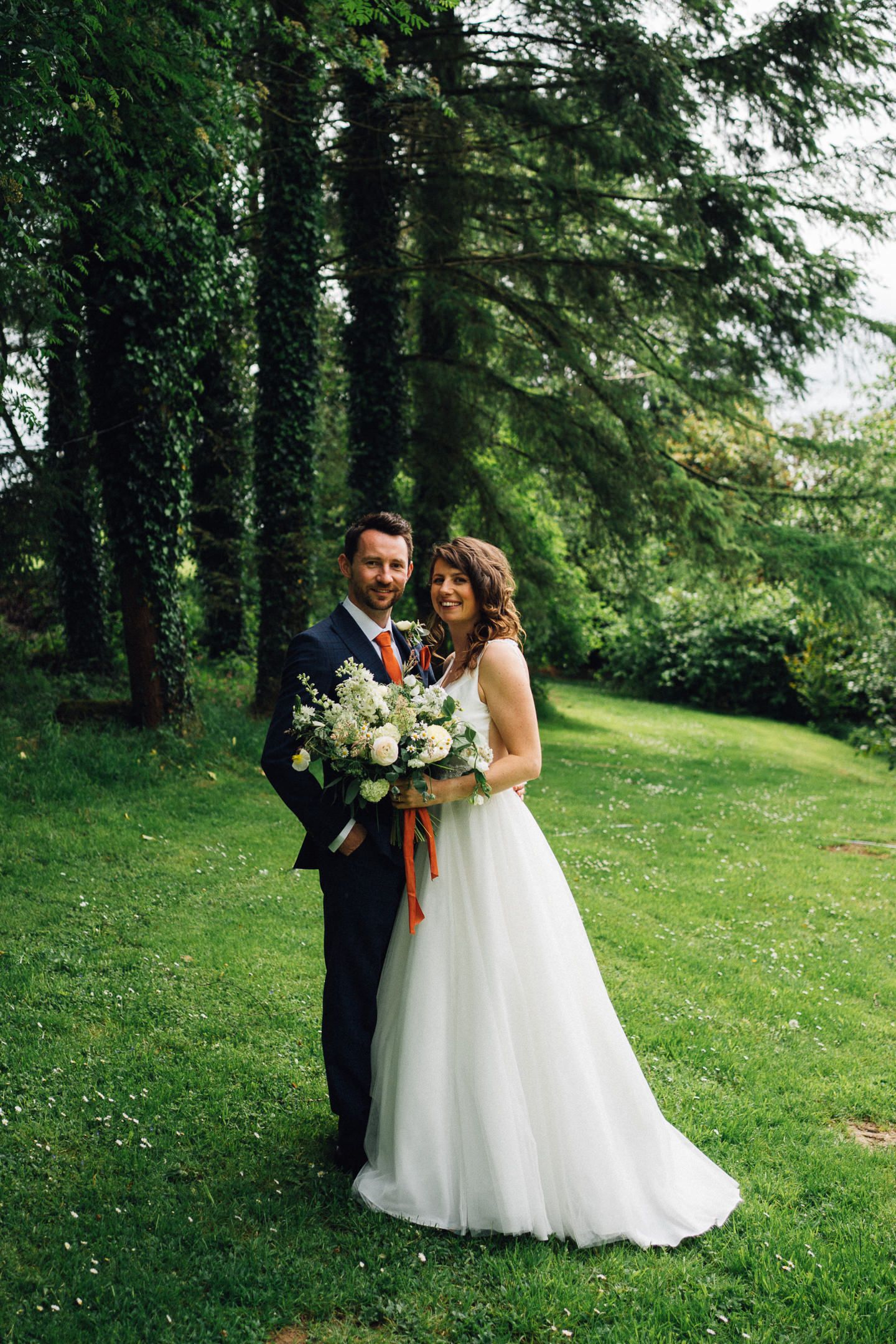 A couple poses together on a lush green lawn surrounded by tall trees.