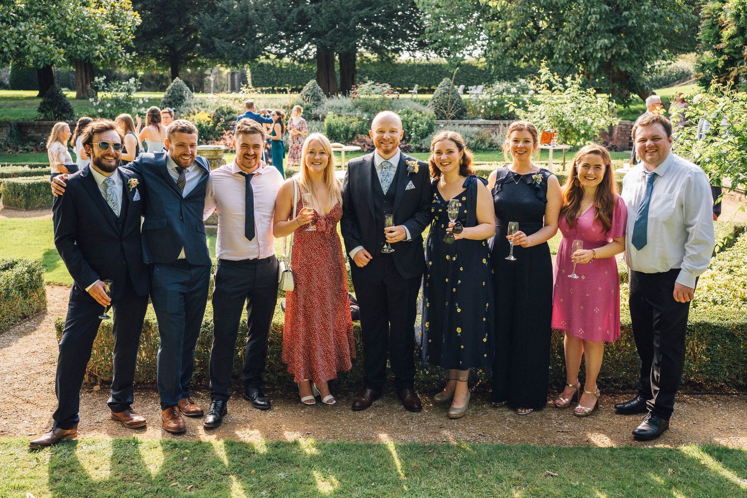Large wedding group photo taken outdoors in a beautiful garden setting.