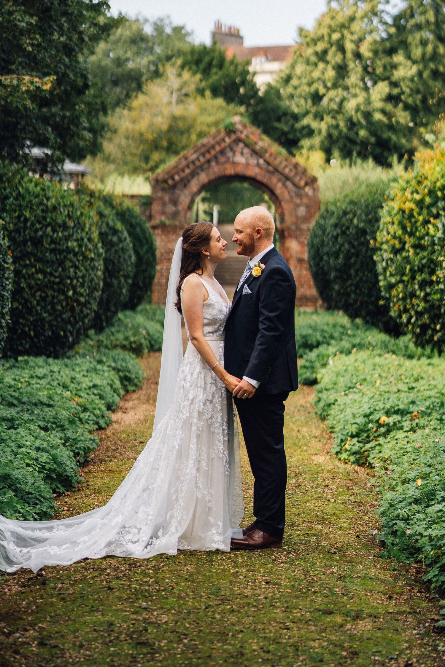 Wedding couple sharing an intimate moment in a garden path lined with manicured hedges.