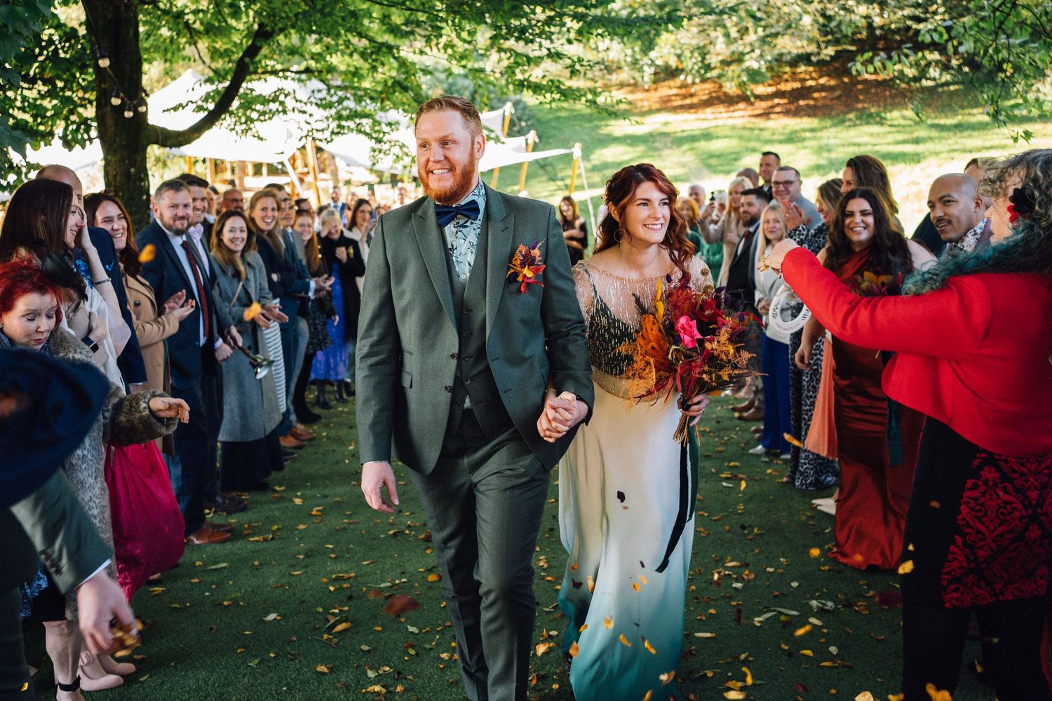 Wedding party walking through a tree-lined path as guests throw autumn leaves in celebration during outdoor ceremony.
