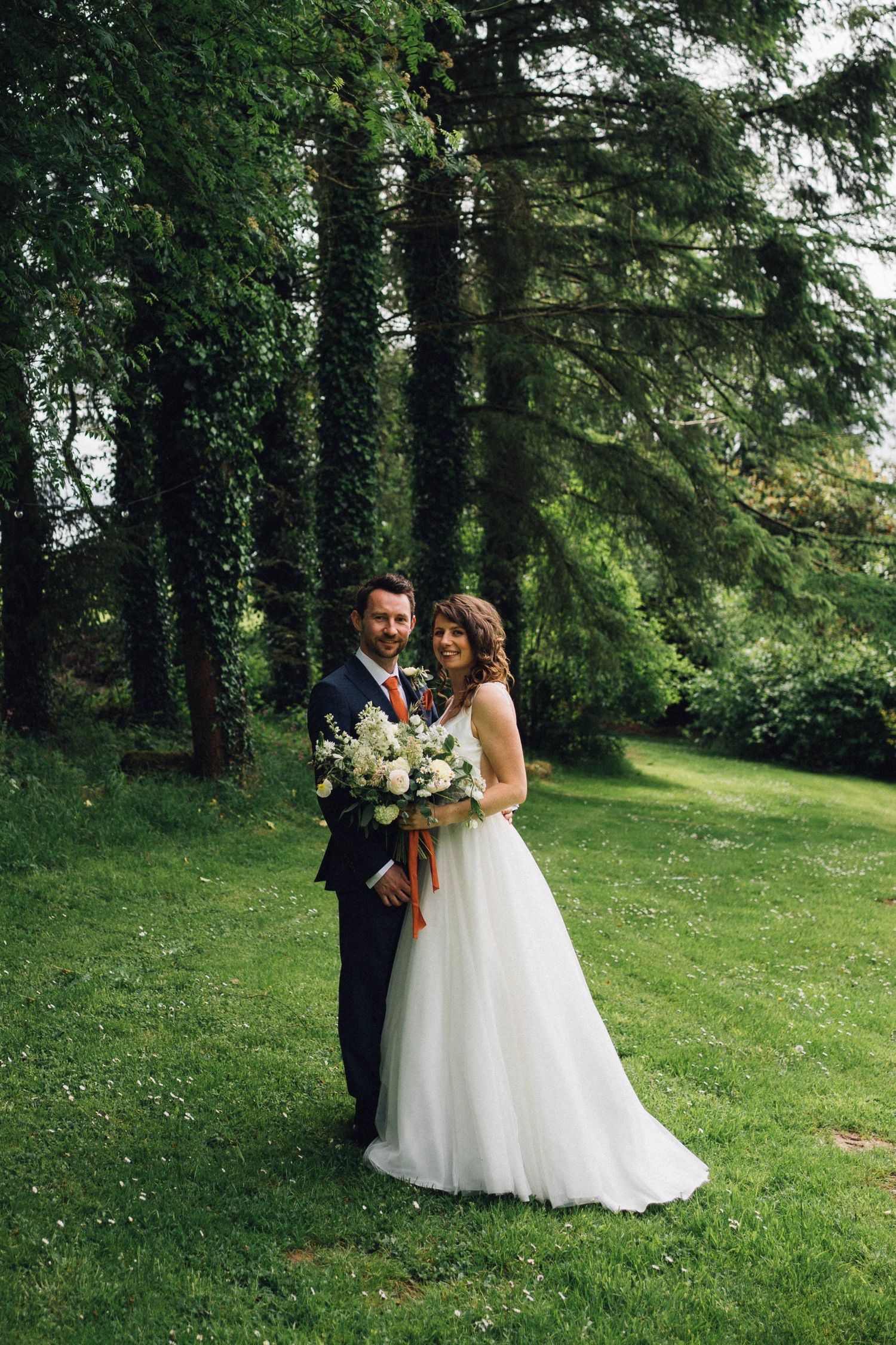 A couple in wedding attire stands together on a lush green lawn surrounded by ivy-covered trees.