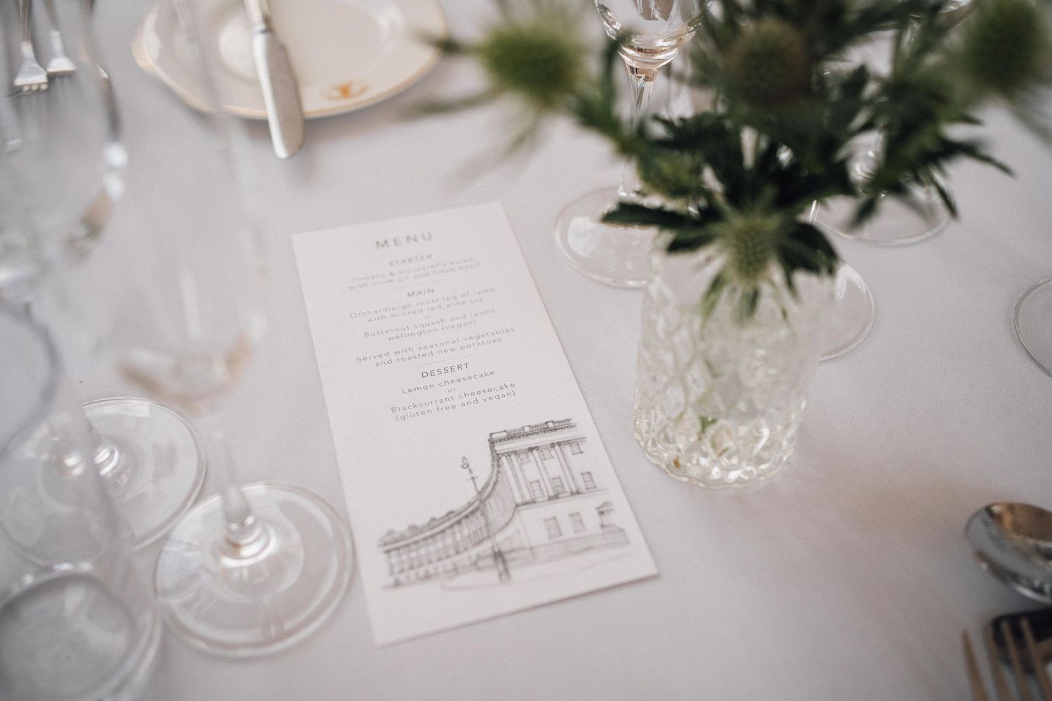 Elegant white wedding reception table setting with menus, glassware and greenery centerpieces.