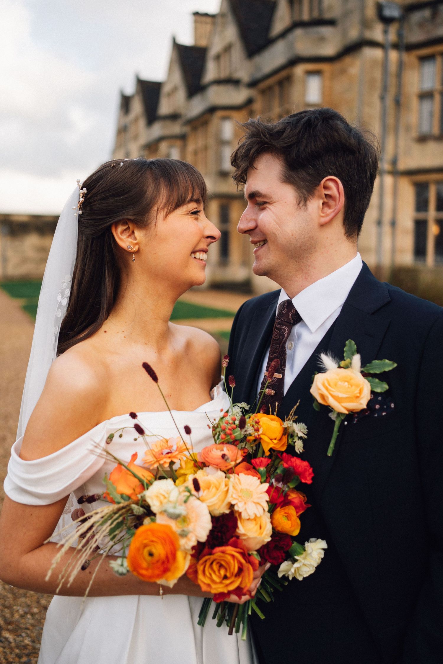 Bride and groom smiling at each other holding a vibrant orange and red bouquet outside a grand stone building.