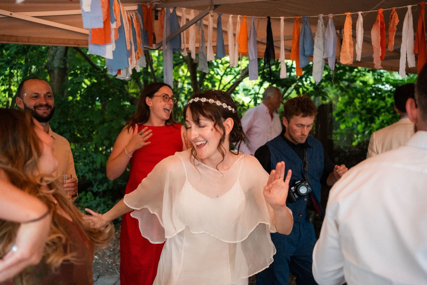 Bride in white dress dancing joyfully with guests at outdoor wedding reception under colorful ribbon decorations.