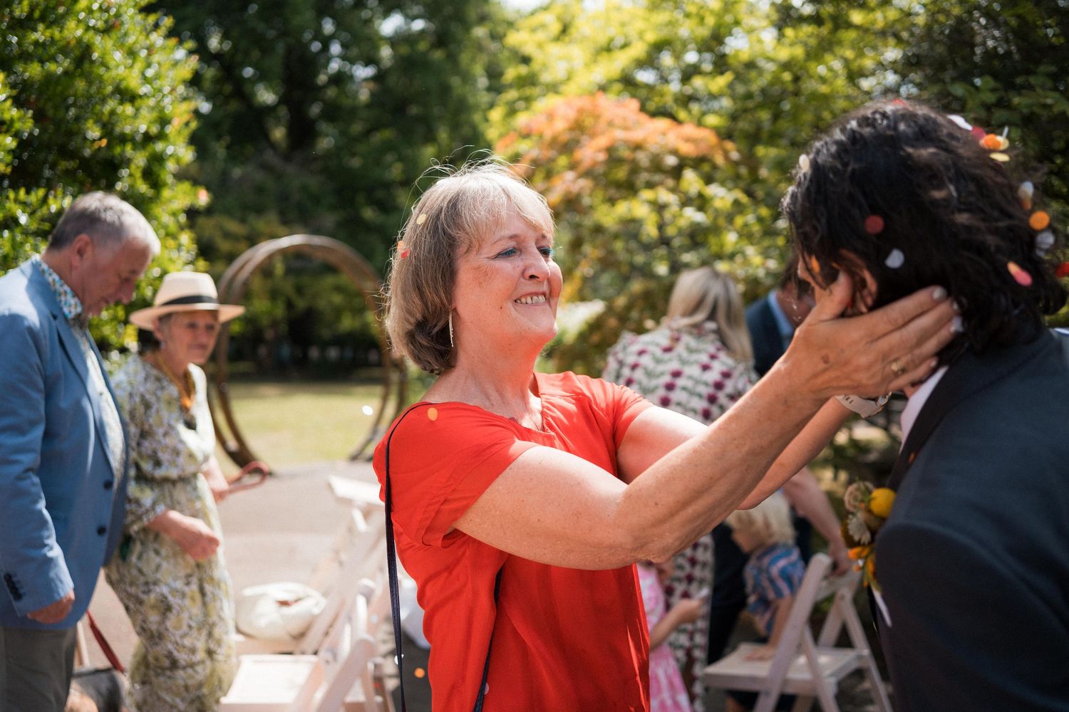 Woman in red top tenderly holds a man's face at an outdoor garden wedding celebration.