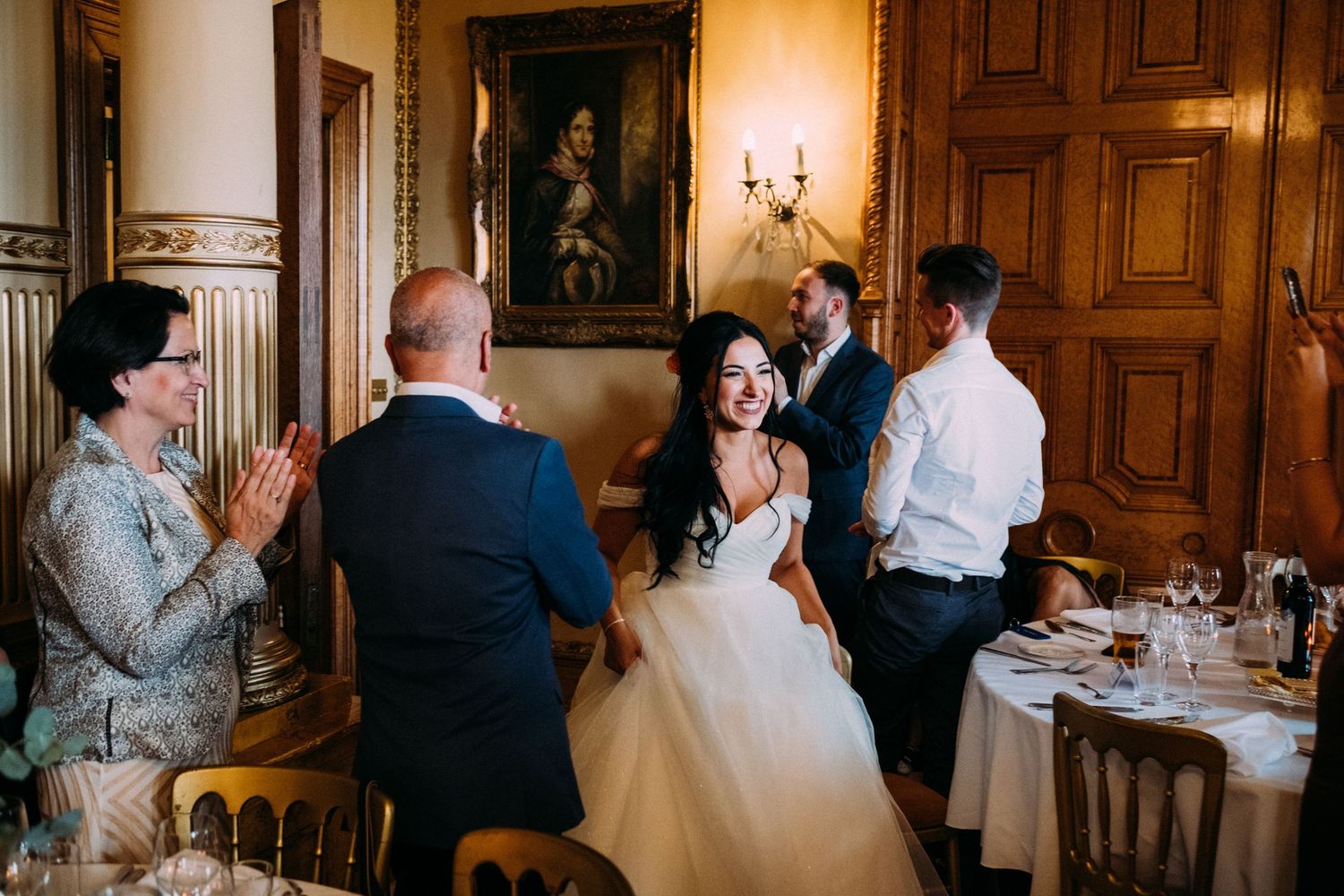 Wedding guests mingle and socialize during an elegant reception in a wood-paneled room with chandeliers.