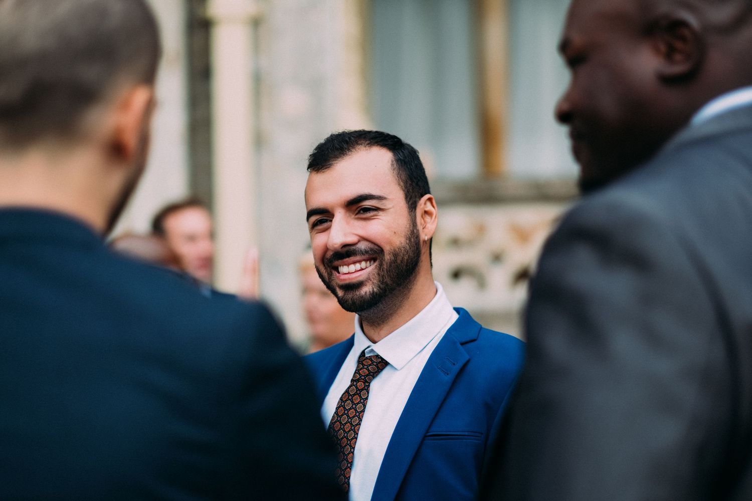 A person in a navy blue suit and tie smiles warmly while engaged in conversation at a gathering.