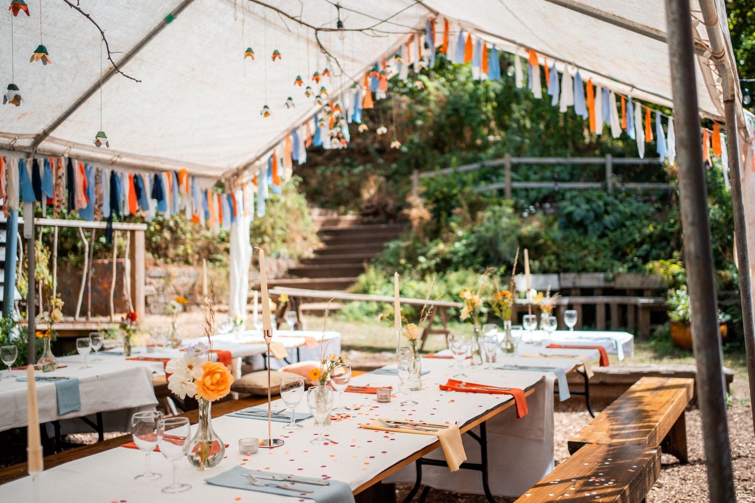 Outdoor wedding reception under a white tent adorned with colorful bunting, set tables with floral centerpieces, and lush garden backdrop.