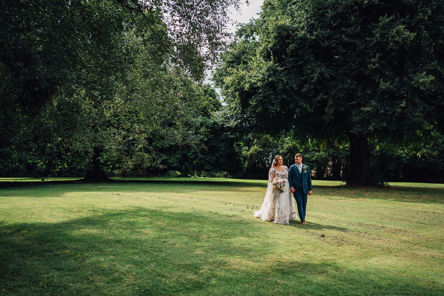 Distant view of newlyweds walking together across a sunlit garden lawn.