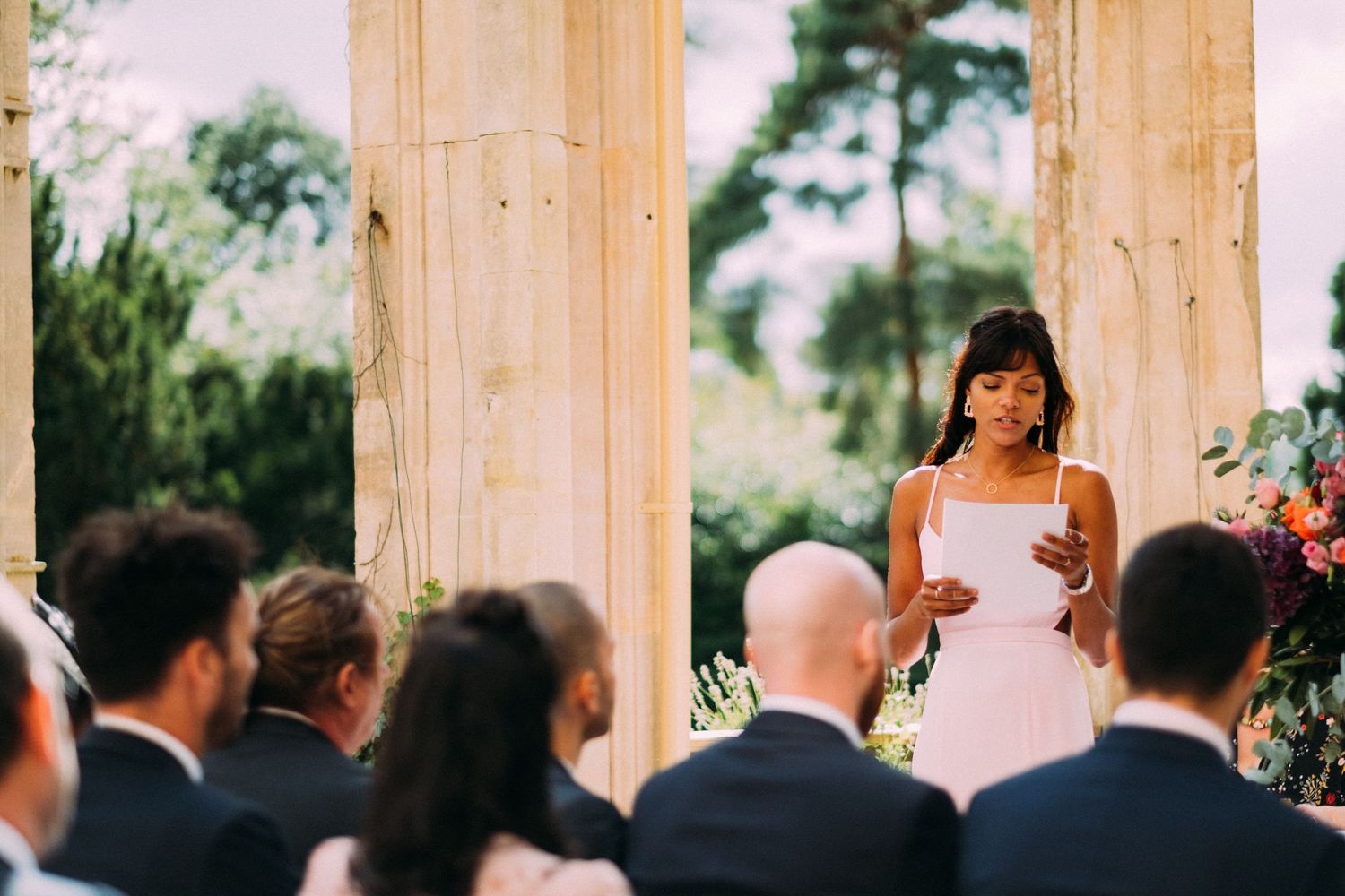 A bride reads her vows during an outdoor wedding ceremony framed by elegant columns.