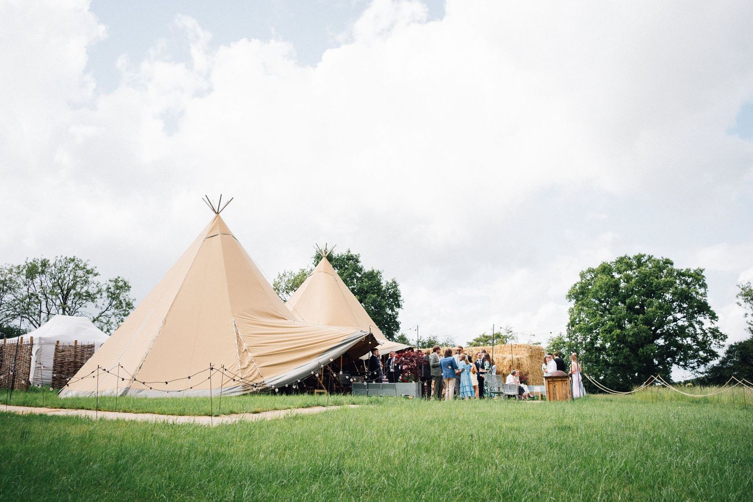 Large beige tipi wedding venue on green grass field with guests gathered outside under a cloudy sky.
