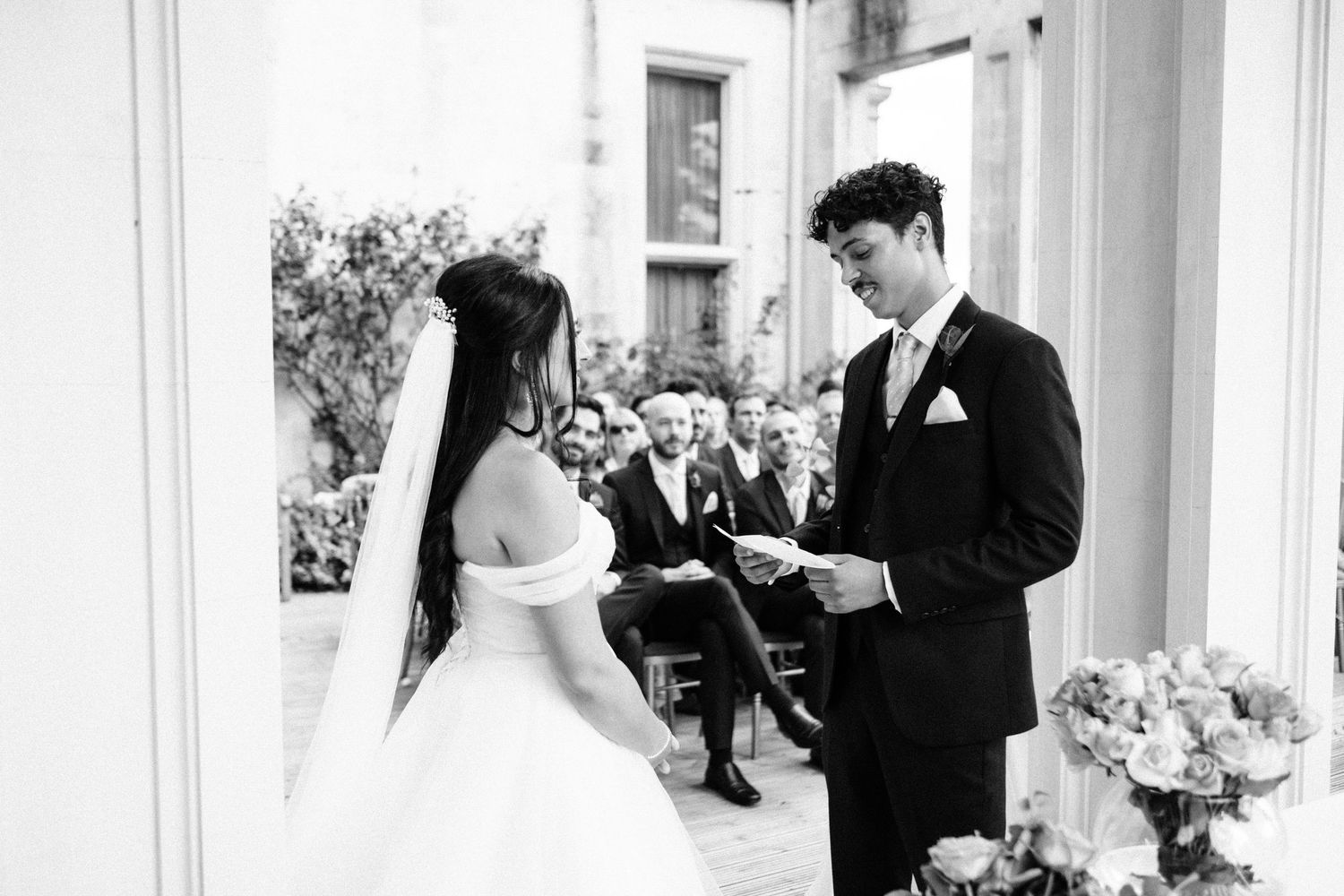 Black and white photo of a groom reading wedding vows to his bride during an outdoor ceremony.