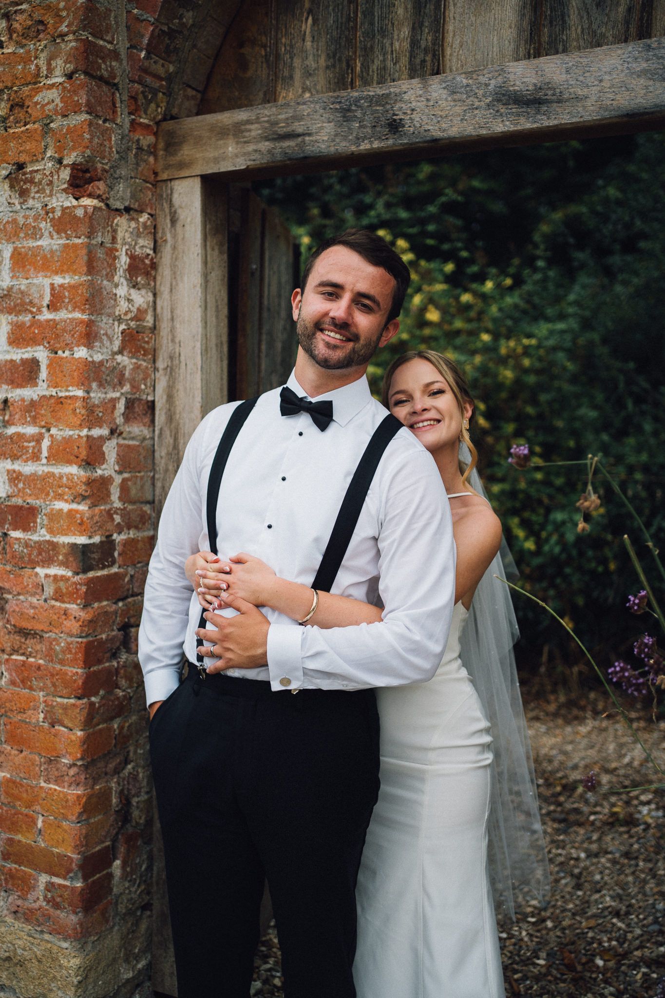 A couple in formal wedding attire poses playfully against a rustic brick wall backdrop.