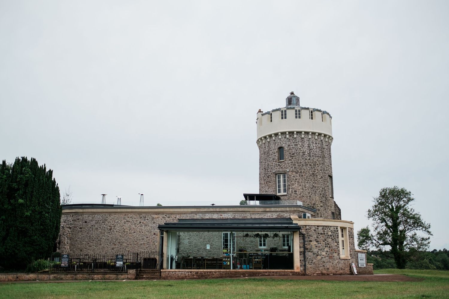 Historic stone observatory tower with circular battlements stands beside a low stone building on a green hill under overcast skies.