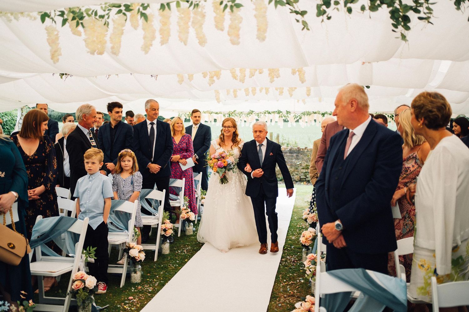 Wedding ceremony guests seated under a white tent decorated with floral arrangements and greenery.