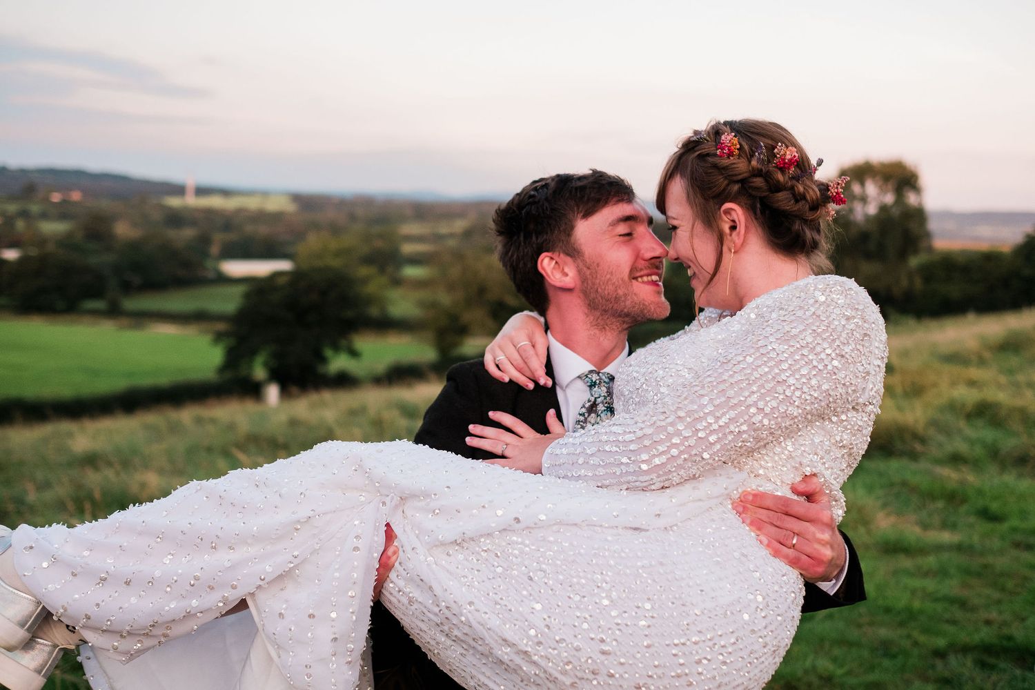 A couple in wedding attire share intimate moments while posing in a scenic countryside setting at sunset.