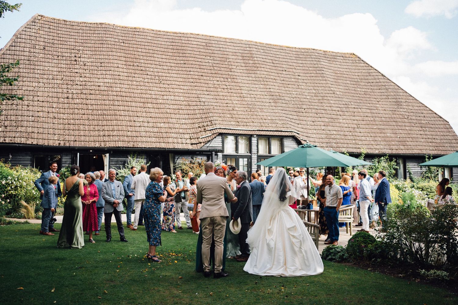 Wedding guests gather outside a rustic barn venue with timber frame architecture and large thatched roof during outdoor reception.