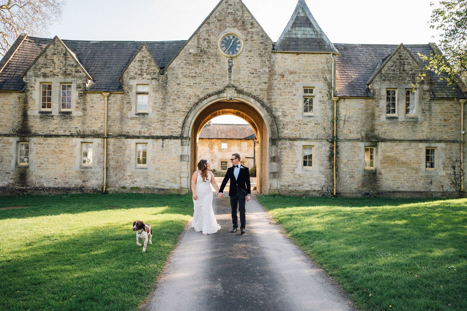 Bride and groom walk hand-in-hand with their dog through a grand stone archway at a historic English countryside wedding venue.