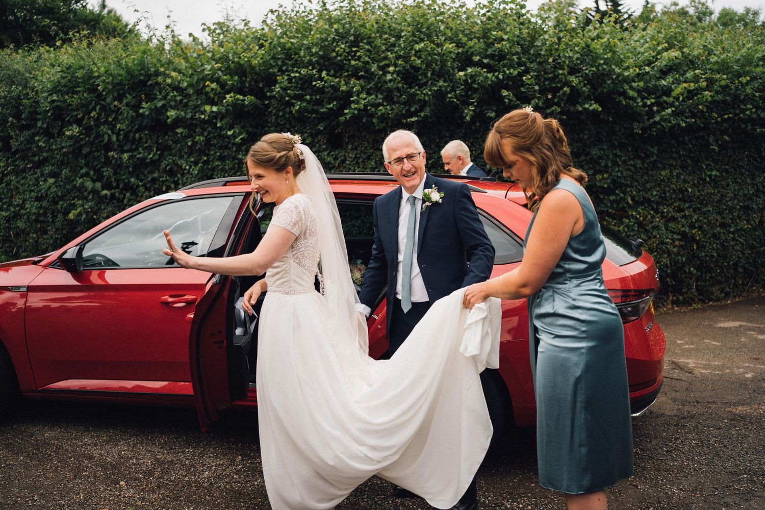 Red car parked outside venue with wedding party nearby.
