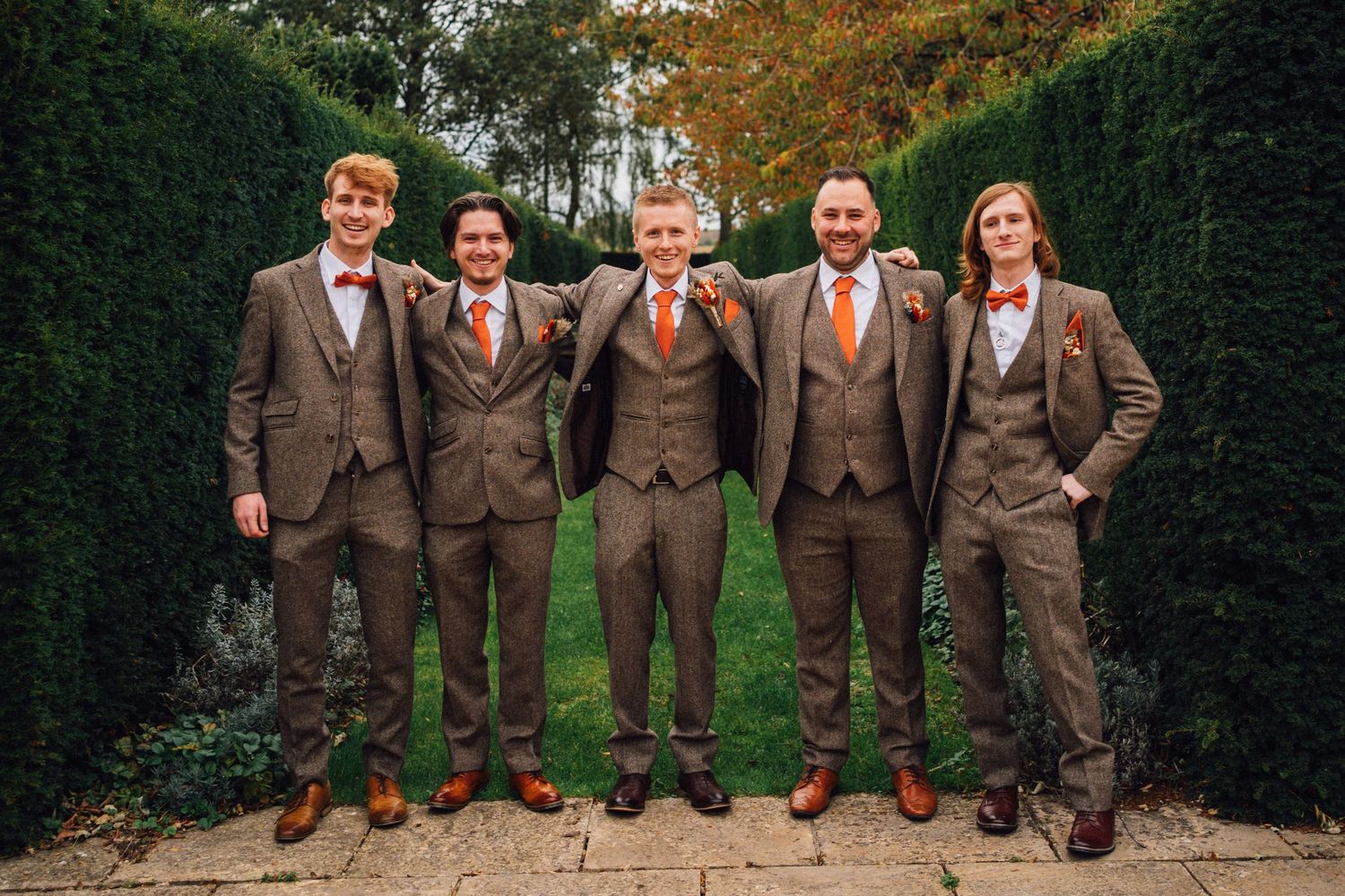 Wedding party wearing matching brown suits and orange ties pose together in an autumn garden setting.