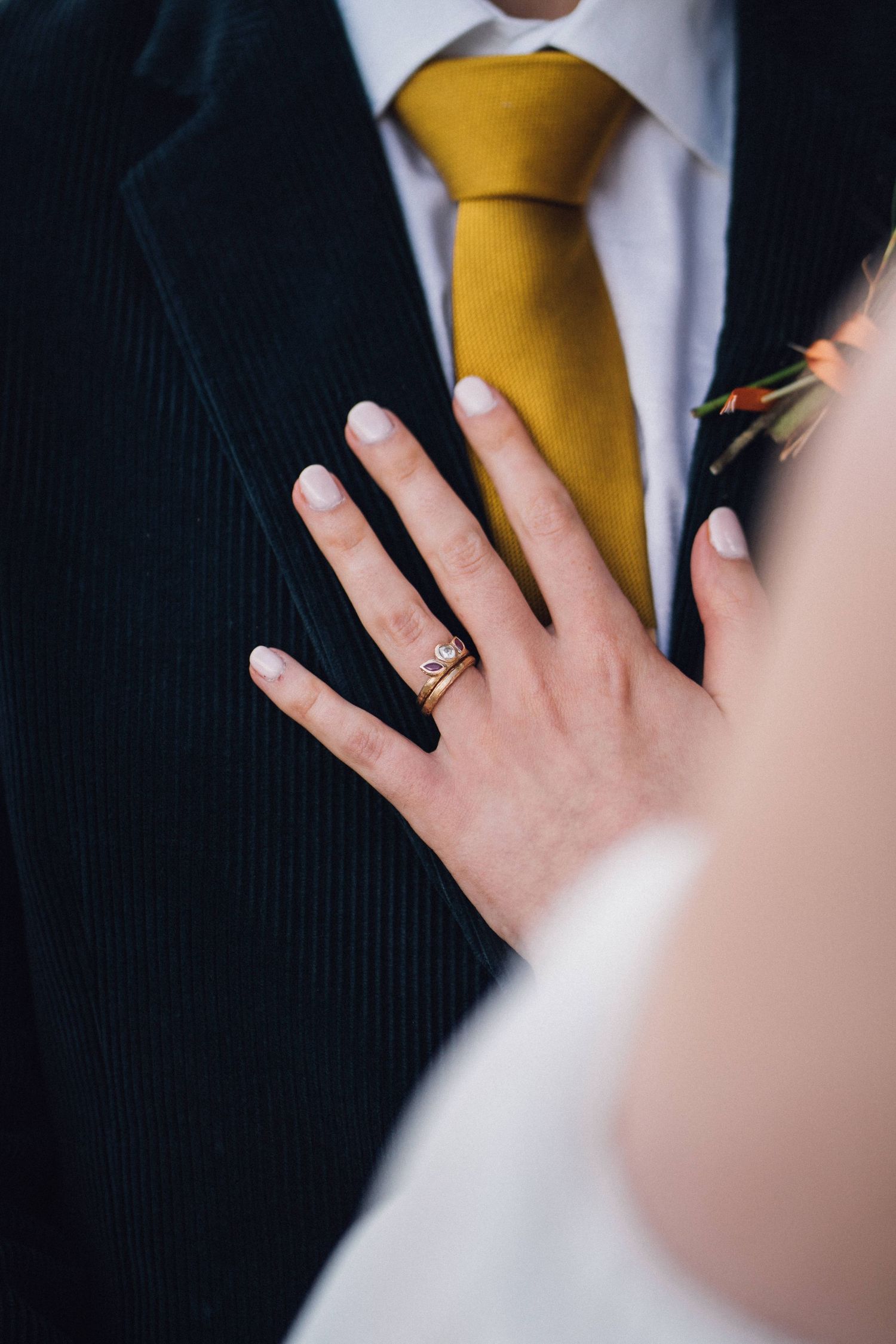 Close-up of hands wearing wedding rings with a yellow tie and dark suit in the background.