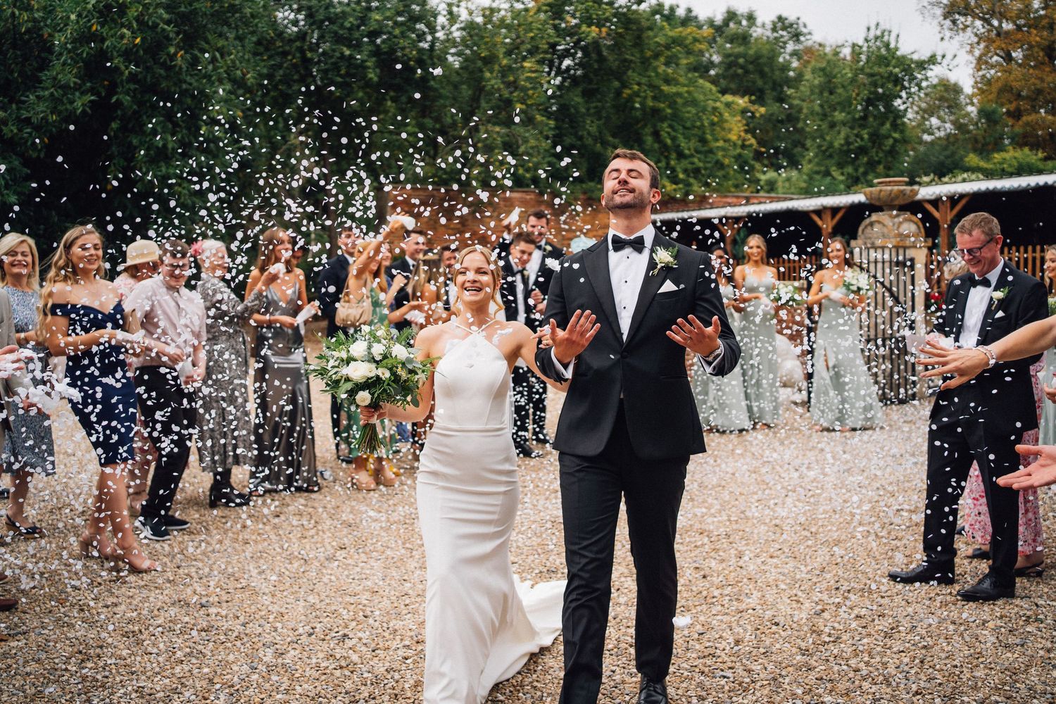 Wedding guests shower the newlyweds with confetti on a sunny outdoor path during the celebration.