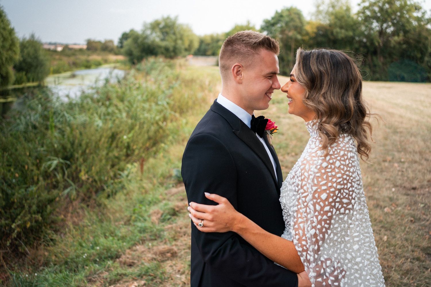 A couple in wedding attire sharing a romantic moment outdoors near a river, groom in black tux, bride in white dress.