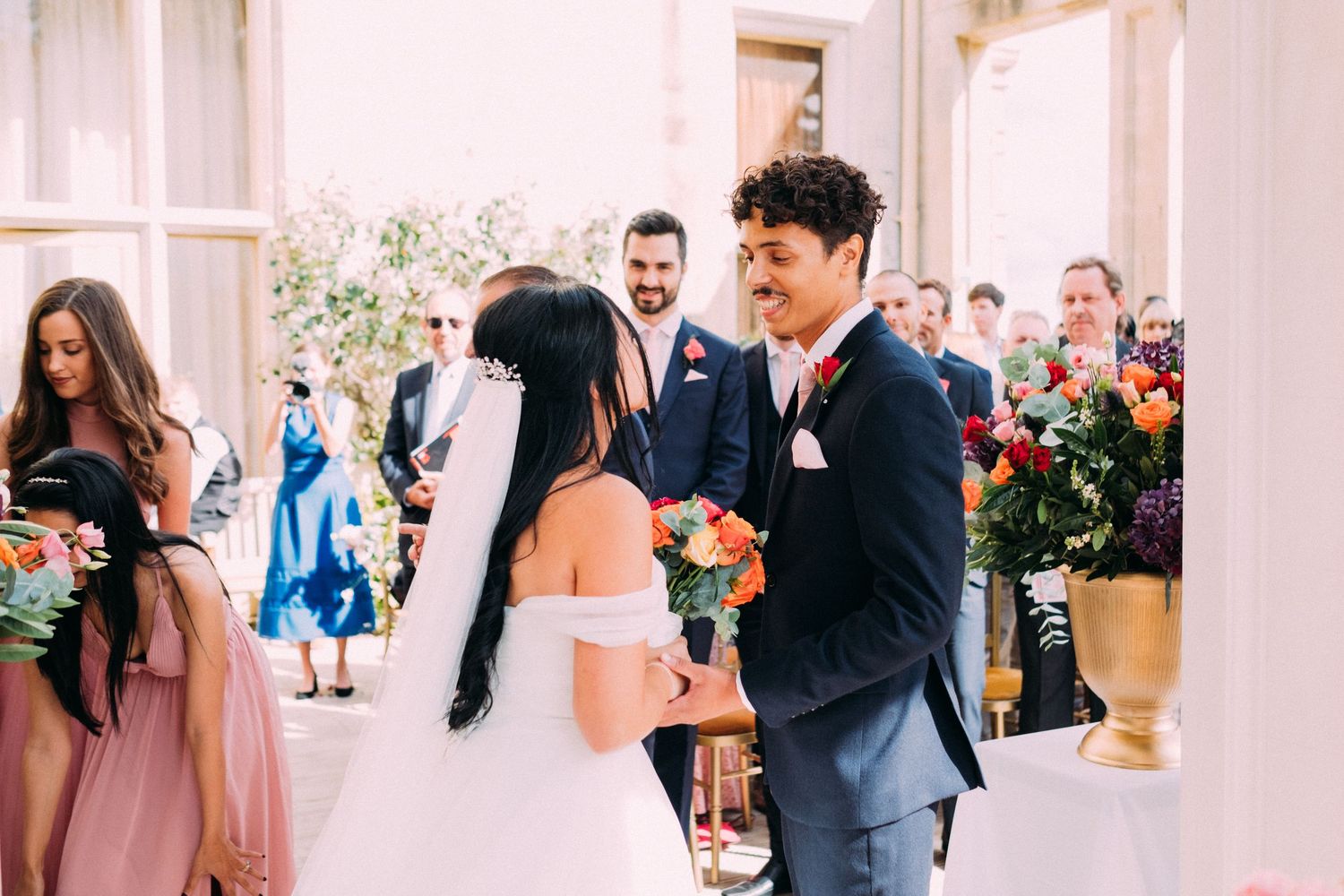 A wedding ceremony takes place in a bright venue with white columns and floral decorations.
