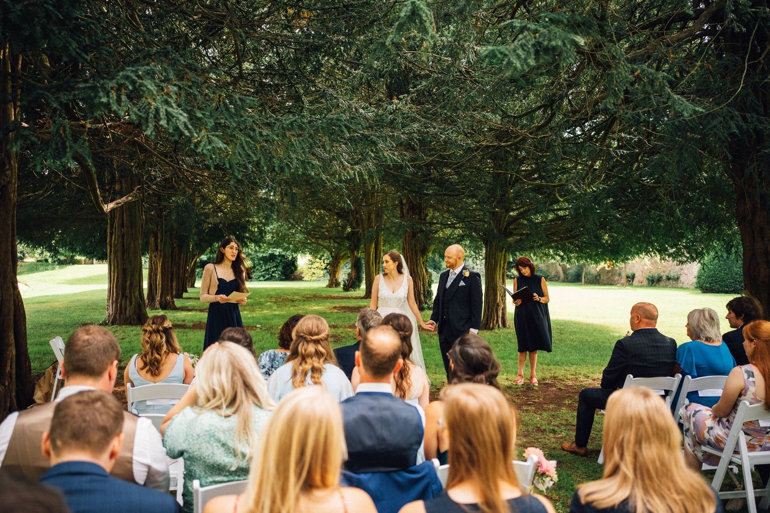 Outdoor wedding ceremony setup with guests seated under large trees.