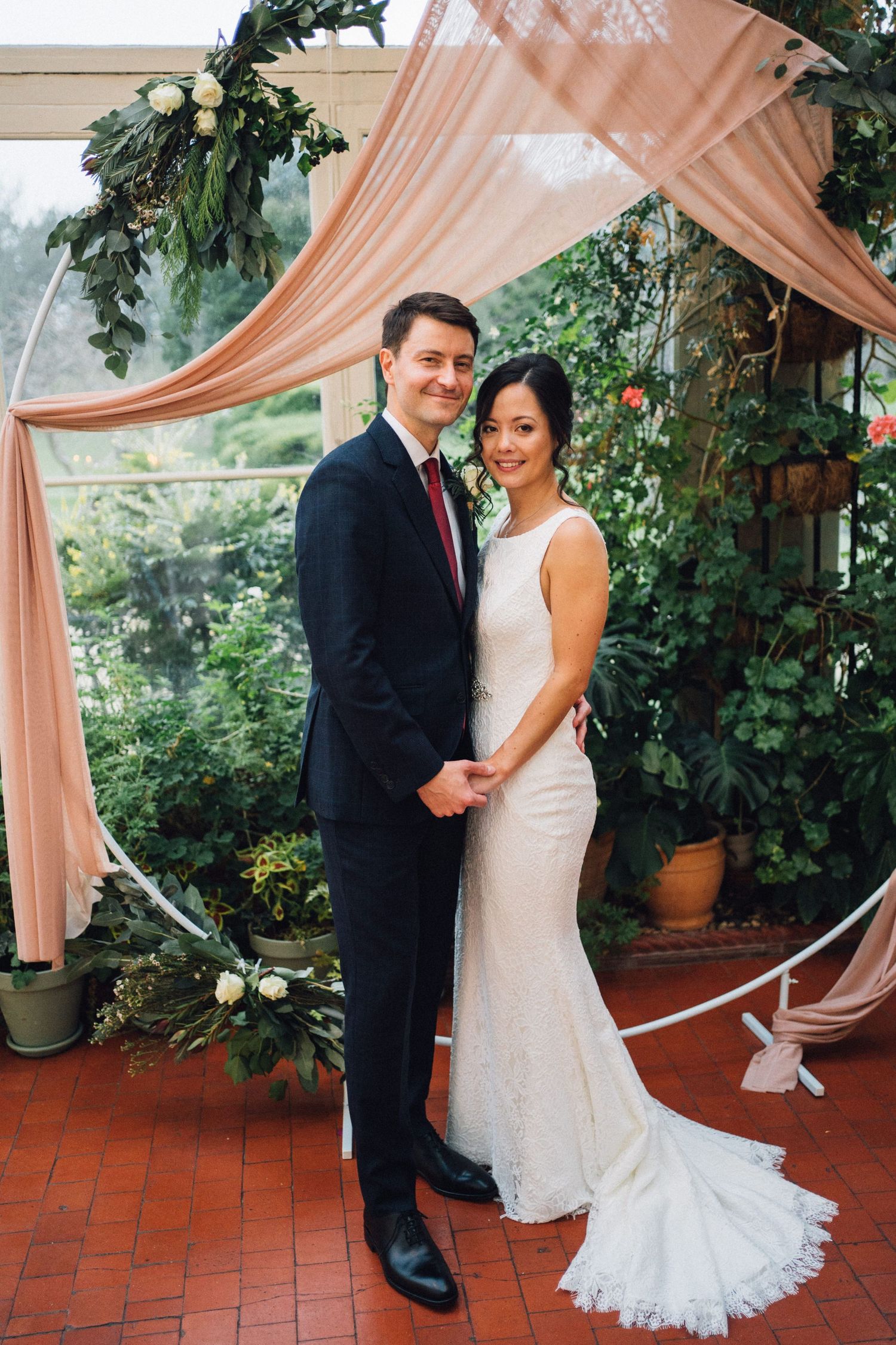 Wedding couple poses under peach drapes and greenery in an outdoor terrace setting.