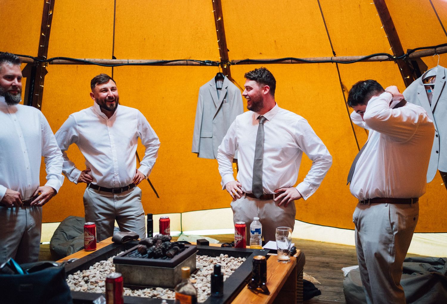 Groomsmen in light colored attire gather inside orange-tinted wedding tent.